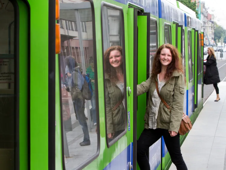Eine junge Frau steigt in eine Stadtbahn der üstra, ihr Gesicht wird in der Seitenscheibe gespiegelt. Am Fahrzeugkopf steigt eine weitere Person ein.