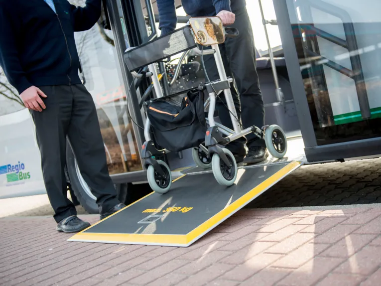 Ein Mann mir einem Rollator, der über eine Rampe aus einem Bus aussteigt. Direkt neben der Tür steht ein weiterer Mann. Von beiden Personen sind lediglich die Beine und ein Teil des Oberkörpers zu sehen.