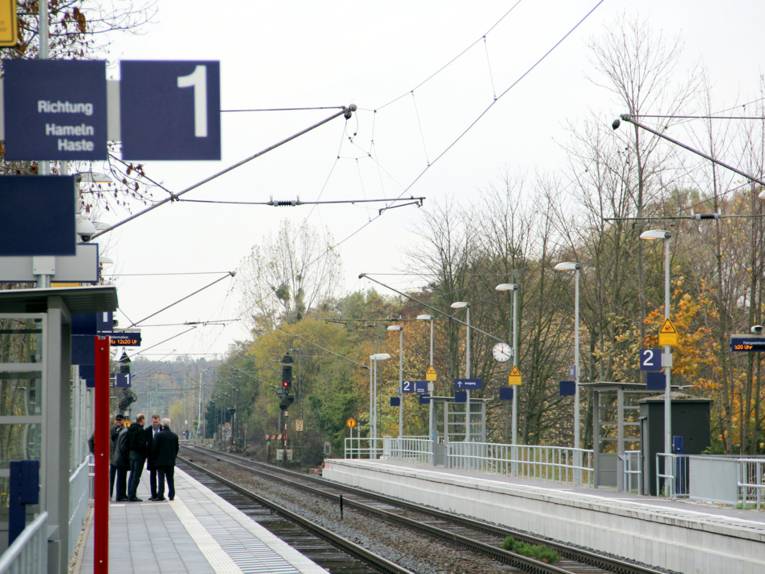 Blick auf den Bahnsteig des S-Bahnhofs Empelde. Links warten Menschen an Bahnsteig 1, rechts leuchtet ein rotes Signal an Bahnsteig 2, dazwischen verlaufen die Schienen.