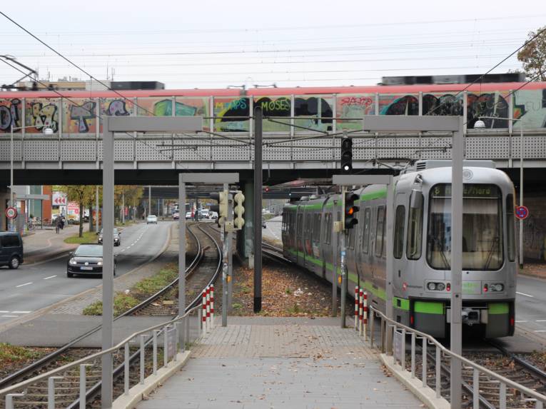 Eine silberne Straßenbahn fährt unter einer Eisenbahnbrücke hindurch. Rechts und links sind mehrspurige Straßen.