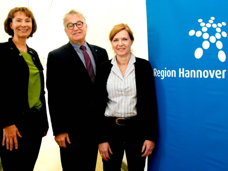 Zwei Frauen und ein Mann neben einem blauen Banner mit Regionslogo.