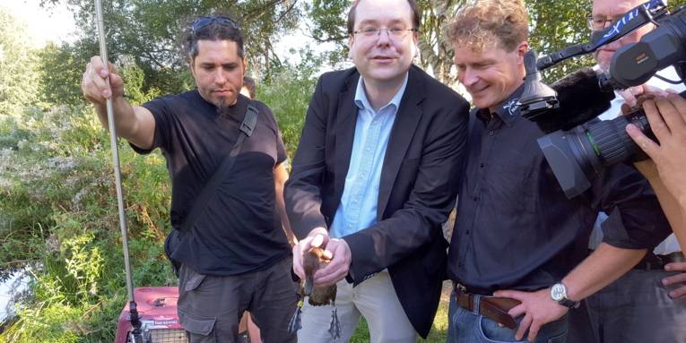 Vier Männer und die Hände eines Kameramanns sind zu sehen. Die zweite Person von links hält eine Moorente.