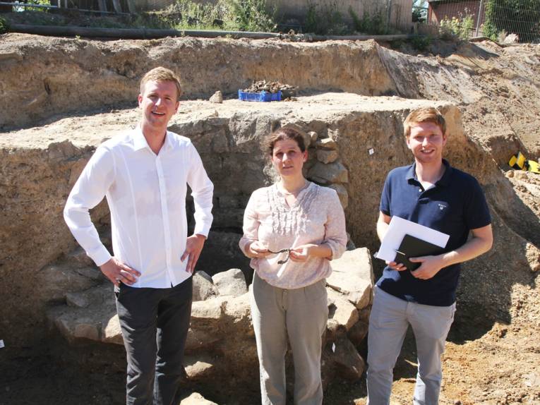 Zwei Männer und eine Frau stehen in einer archäologischen Grabungsstelle.