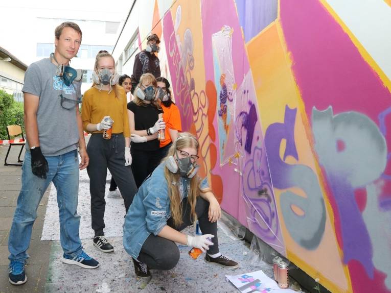 Sechs Jugendliche und ein Erwachsener gestalten eine Wand mit Graffitikunst.