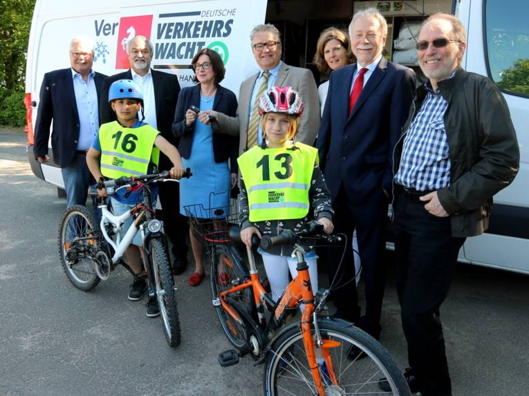 Fünf Männer und zwei Frauen stehen vor einem weißen Kleintransporter, davor sind ein Junge und ein Mädchen samt ihren Fahrrädern, Fahrradhelmen und nummerierten Warnwesten.