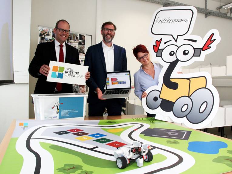 Eine Frau mit einem Aufsteller mit der Aufschrift "Willkommen", ein Mann mit einem Laptop und ein weiterer Mann mit einem Schild, auf dem "Open Roberta Coding Hub" steht.