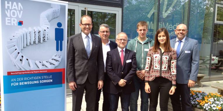 Sechs Personen haben sich zu einem Foto zusammengestellt, daneben steht ein Rollup-Display mit dem Text "Beschäftigungsförderung Region Hannover. An der richtigen Stelle für Bewegung sorgen"