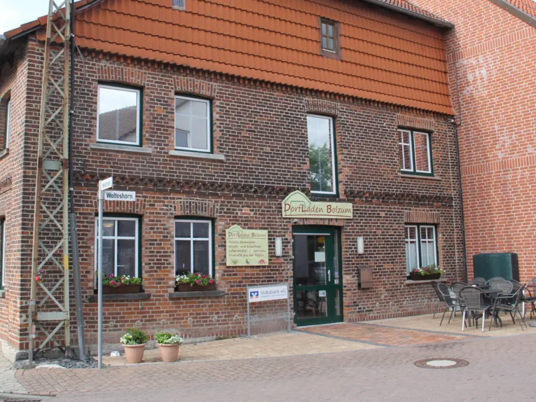 Eingang in ein Backsteingebäude, davor Blumenkübel und ein Tisch mit Korbstühlen