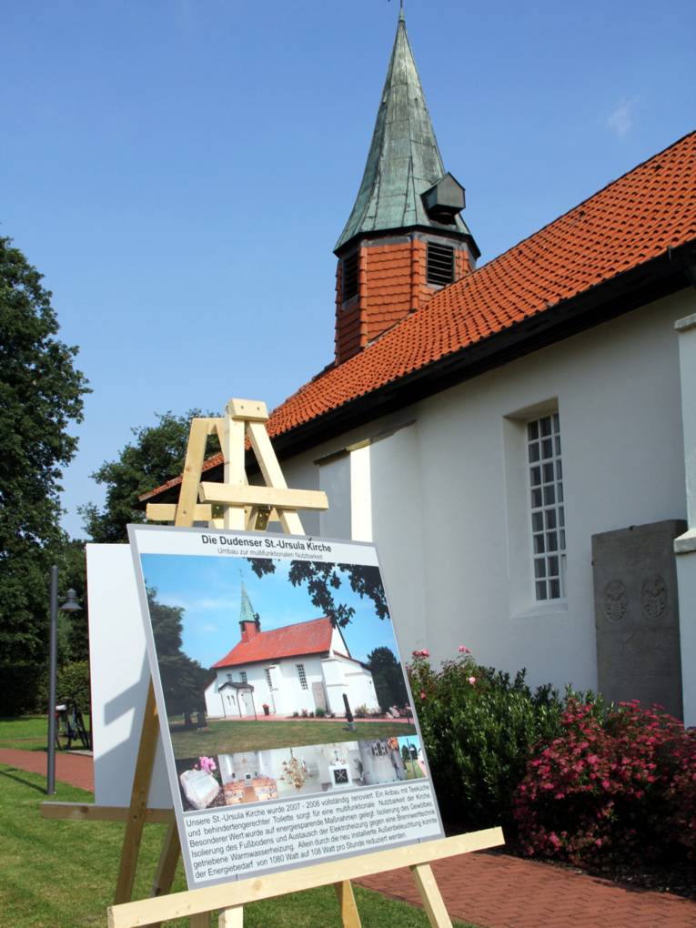 Kirchengebäude mit weißer Fassade, rotem Ziegeldach und kleinem Turm. Davor steht eine Infotafel auf einer Staffelei.