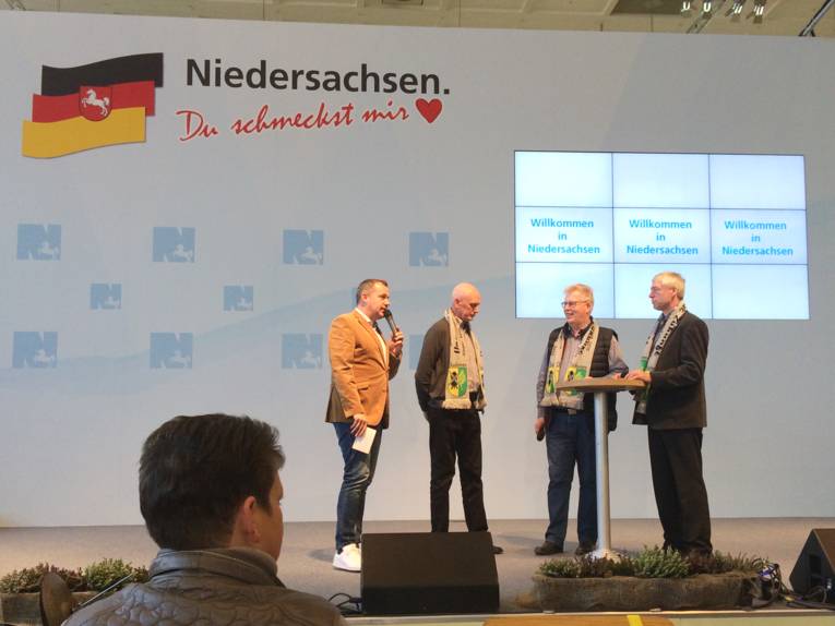 Vier Männer stehen auf einer Bühne. Im Hintergrund steht auf einer Wand "Niedersachsen. Du schmeckst mir. Drei Monitore zeigen außerdem jeweils "Willkommen in Niedersachsen".