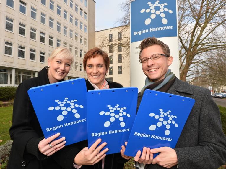 Zwei Frauen und ein Mann stehen draußen nebeneinander und halten jeweils ein Klemmbrett in die Kamera.