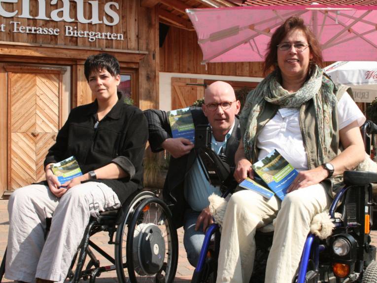 Links eine Frau im Rollstuhl. Rechts eine Frau im Rollstuhl. In der Mitte ein Mann der zwiscchen den beiden Frauen in die Hocke gegangen ist.