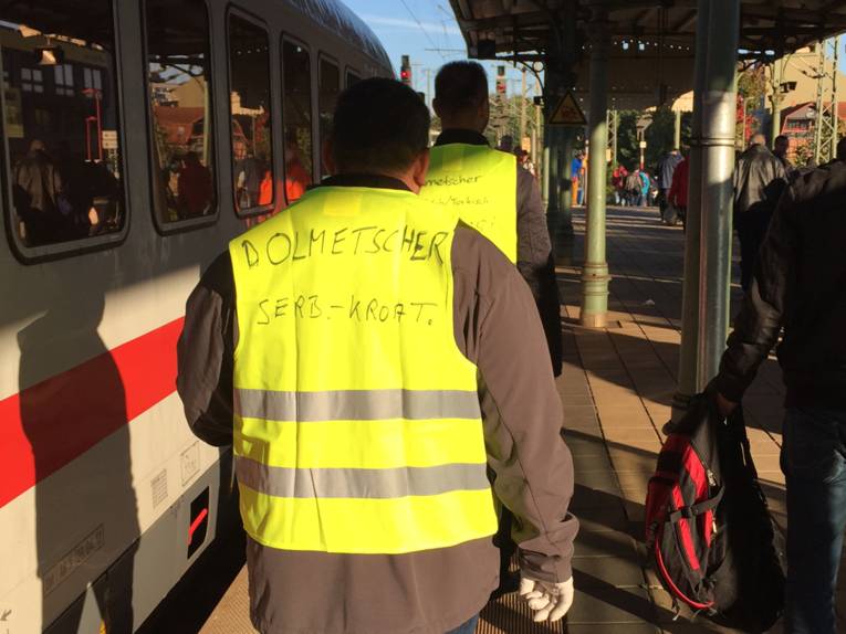 Zwei Männer gehen einen Bahnsteig entlang. Sie tragen Warnwesten, auf denen mit Filzstift "Dolmetscher" geschrieben steht und welche Sprachen sie beherrschen. Links neben ihnen steht ein Zug im Gleis.