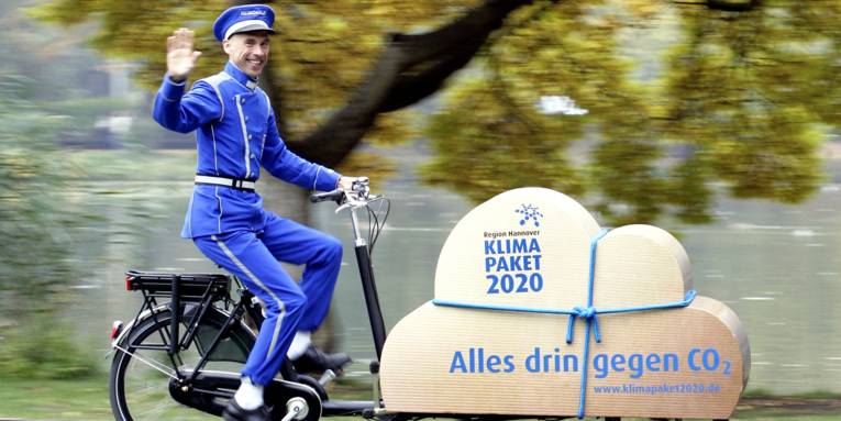 Ein Mann trägt eine Dienstbotenuniform und fährt auf einem Lastenfahrrad. Auf der Ladefläche des Fahrrads ist eine braune Wolke in Paketform. Darauf steht "Region Hannover. Klimapaket 2020. Alles drin gegen CO2 – www.klimapaket2020.de