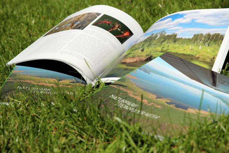 Ein Buch liegt aufgeschlagen auf grünem Rasen, zwei weitere Exemplare liegen mit dem Titel nach oben daneben. Die Seiten zeigen links Text mit zwei Bildern, rechts ein seitenfüllendes Foto einer Moorlandschaft.