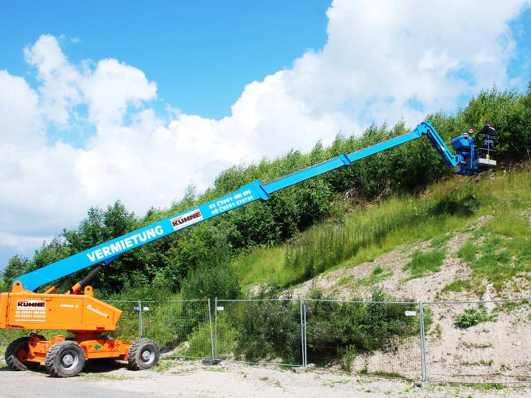 Kran mit ausgefahrenem Ausleger, der Ausleger reicht über eine umzäunte, bewachsene Halde