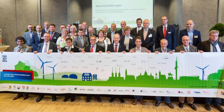 Gruppenfoto mit vielen Personen. Die erste Reihe hält ein sehr breites Banner.