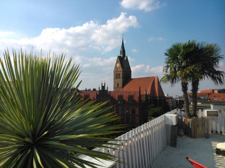 Blick von einer mit Sand und Palmen dekorierten und einem weißen Holzzaun abgegrenzten Dachterrasse über die Dächer der Innenstadt Hannovers auf die Marktkirche 