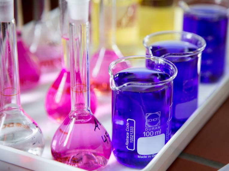 Kolbengefäße mit schmalem Hals (links) und zylindrische Becher mit Ausguss (rechts). Die enthaltenen Flüssigkeiten sind in den Farben Rosa (Messkolben) und Blau (Bechergläser) eingefärbt. 