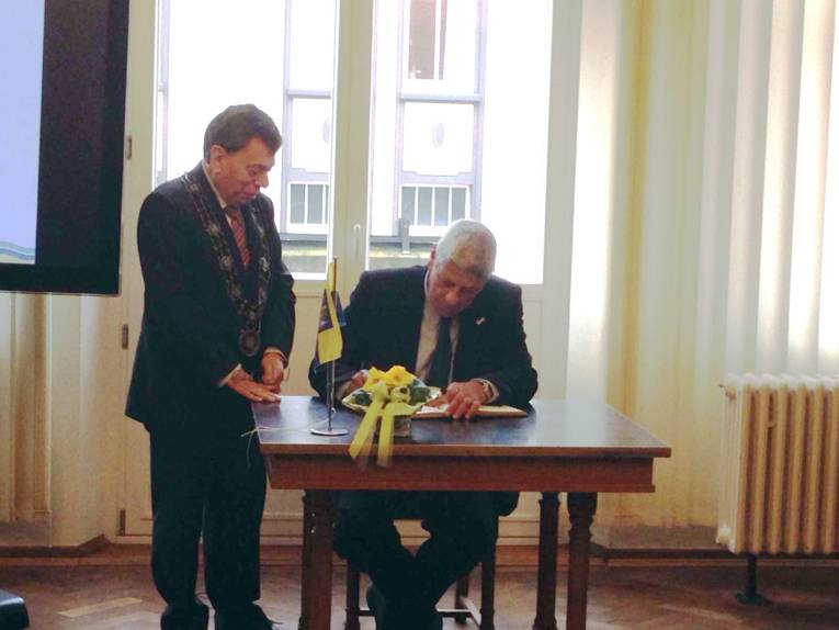 Moti Dotan sitzt an einem Tisch und schreibt, daneben steht Rolf-Axel Eberhardt, der seine Bürgermeisterkette trägt.
