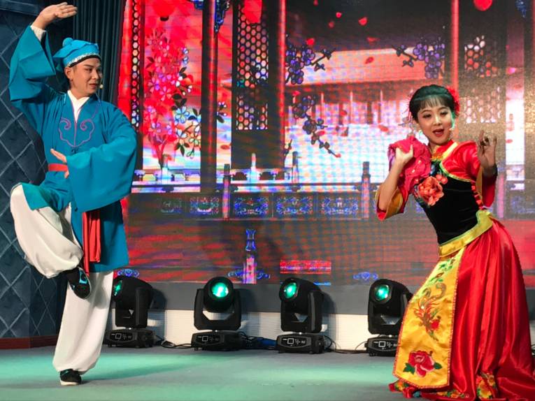 Ein Mann und eine Frau stehen in farbenfrohen Gewändern auf einer Bühne und tragen etwas vor.