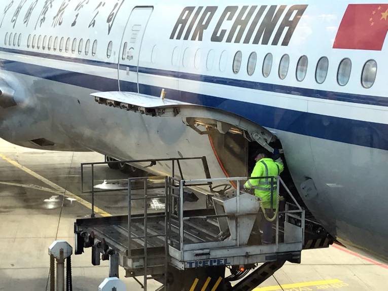 Ein Arbeiter steht auf einer mobilen Hebebühne an der geöffneten Frachtraumluke eines Verkehrsfluigzeugs. Auf dem Flugzeug sind chinesische Schriftzeichen und die Buchstaben "AIR CHINA".
