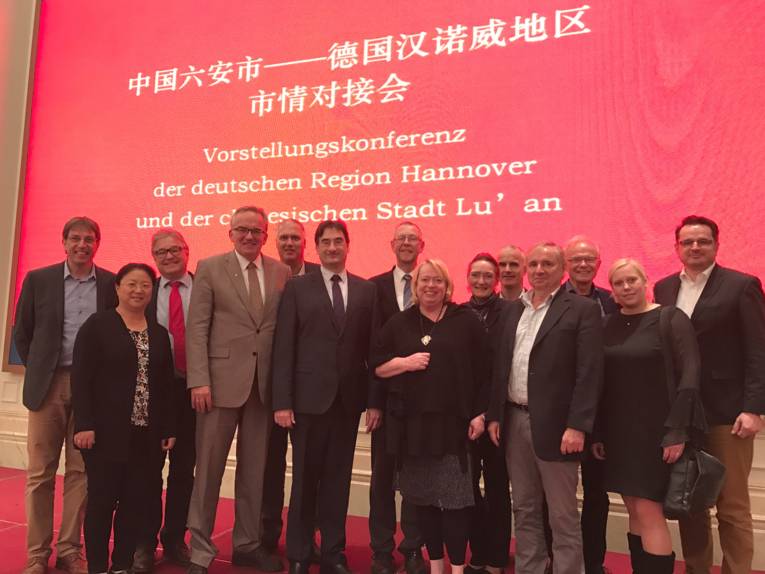 Vierzehn Personen stehen vor einer roten Leindwand, oben stehen chinesische Schriftzeichen, darunter der Text "Vorstellungskonferenz der deutschen Region Hannover und der chinesischen Stadt Lu´ an 