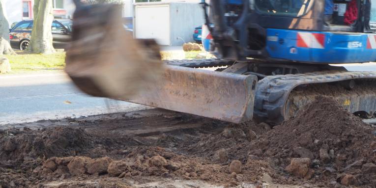 Ein blauer Bagger hebt ein Loch aus. Die Schaufel bewegt sich schnell und wirkt dadurch dynamisch unscharf.