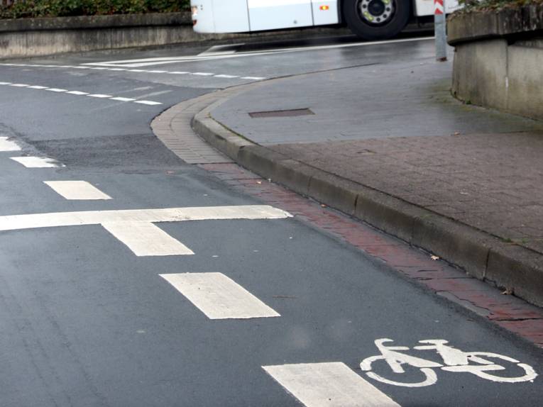 Eine weiße, gestrichelte Linie trennt auf einer Straße einen Bereich für Radfahrende ab. Im geschützten Bereich ist ein Fahrrad-Piktogramm, vor einer Kreuzung ist eine Haltelinie.
