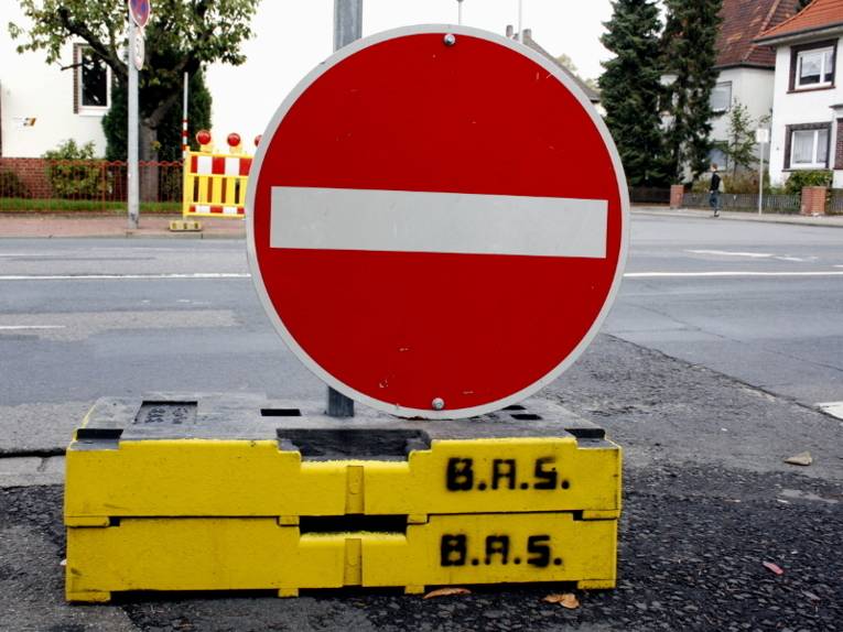 Ein Verkehrsschild "Verbot der Einfahrt" wird provisorisch von schweren Betonelementen gehalten. Im Hintergrund ist eine Straßenkreuzung zu erkennen.