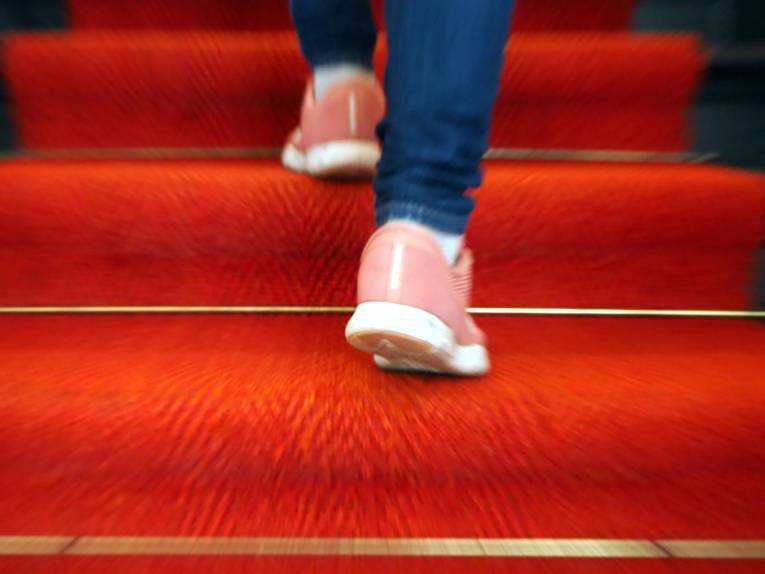 Zwei Füße stecken in Turnschuhen und bewegen sich schnell eine Treppe hinauf. Die Treppe ist mit rotem Teppich ausgelegt.