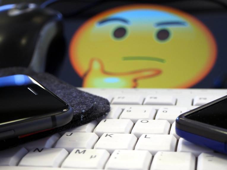 Auf einer Tastatur liegen zwei Smartphones, von einem Tablett dahinter strahlt ein grübelnder Smiley.