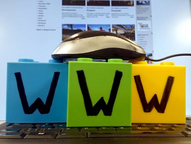 Eine Maus liegt auf drei Plastikbausteinen, diese wiederum stehen auf einer Tastatur, dahinter zeigt ein Bildschirm eine Webseite an. Auf jedem der drei Bausteine steht der Buchstabe "W".