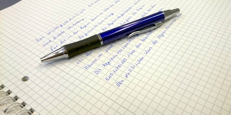 Das oberste Blatt eines Schreibblocks ist beschrieben. Obenauf liegt ein blauer Kugelschreiber.