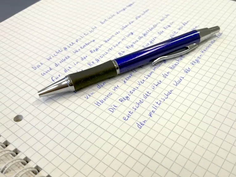 Das oberste Blatt eines Schreibblocks ist beschrieben. Obenauf liegt ein blauer Kugelschreiber.