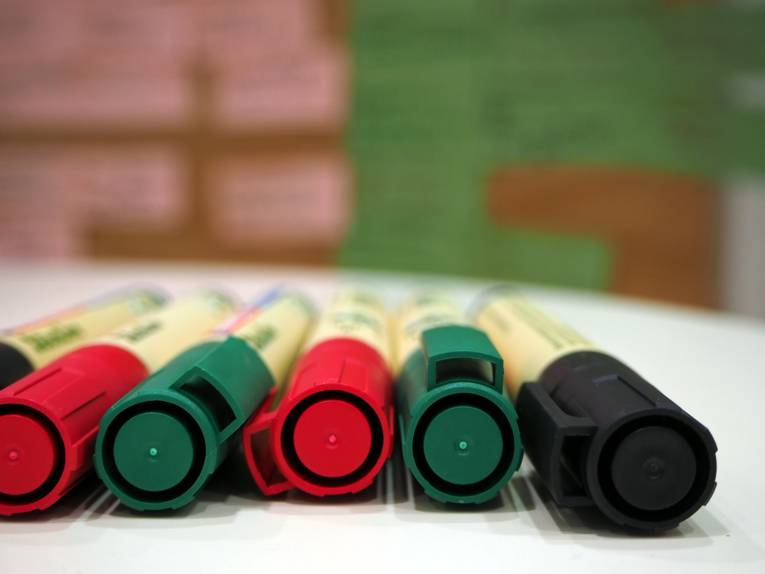 Filzstifte liegen nebeneinander auf einem Stehtisch, im Hintergrund ist eine Metaplanwand mit Kärtchen zu sehen.