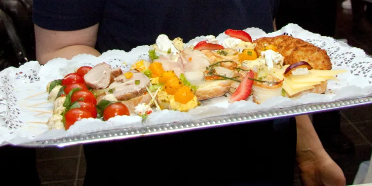 Tomaten und Mozzarella, Braten, Erdbeeren und andere Leckereien liegen auf einem Tablett, das eine Person mit einer Hand trägt.