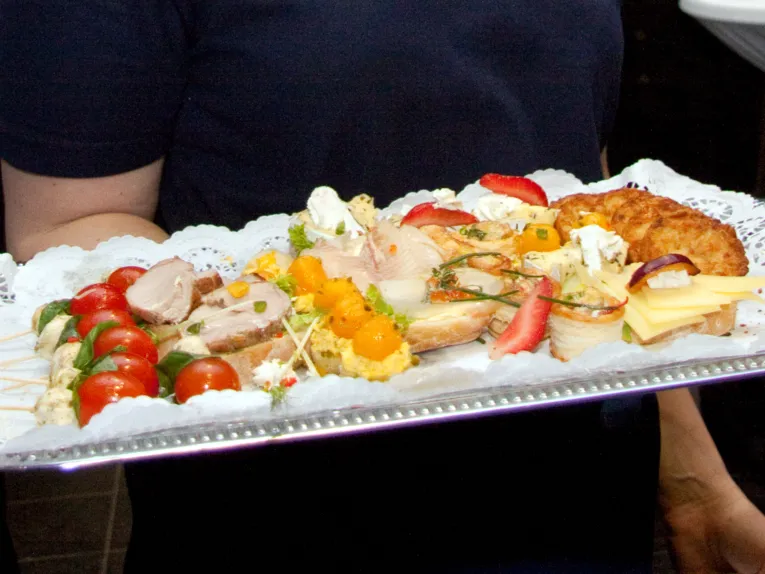 Tomaten und Mozzarella, Braten, Erdbeeren und andere Leckereien liegen auf einem Tablett, das eine Person mit einer Hand trägt. 