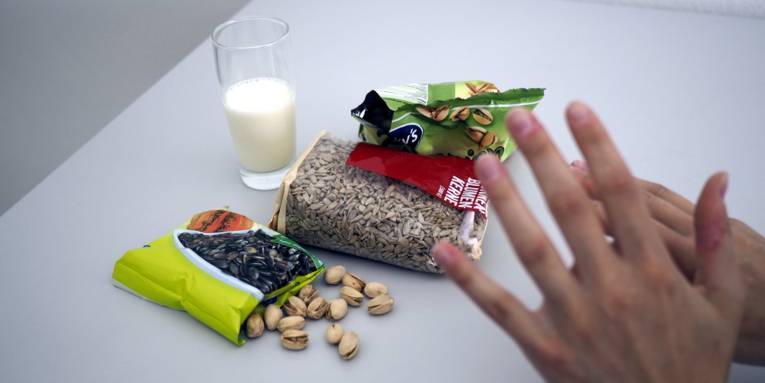 Ein Glas Milch, eine Packung Kürbiskerne, eine Packung Sonnenblumenkerne, eine Packung Pistazien und einzelne Pistazien auf befinden sichnauf einem Tisch. Über den Lebensmitteln sind zwei Hände zu sehen, mit denen eine ablehnende Haltung gezeigt wird.