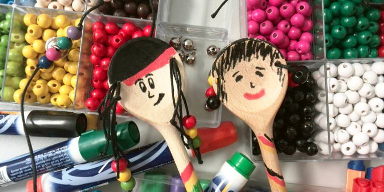 Zwei Kochlöffel aus Holz wurden angemalt und mut Bändern und Holzperlen ausgestattet. Im Hintergrund ist weiteres Bastelmaterial, zum Beispiel weitere Holzperlen in verschiedenen Farben und Glöckchen aus Metall.