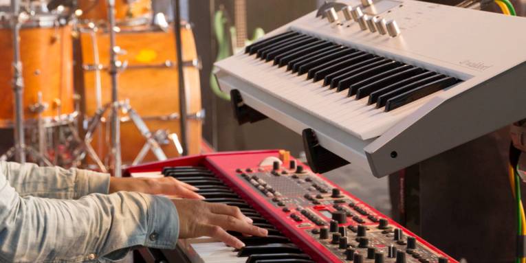 Hände bespielen ein Keyboard, im Hintergrund sind weitere Musikinstrumente.