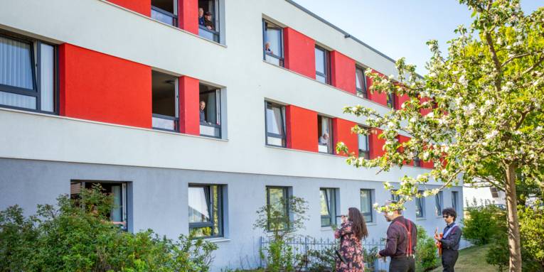 Zwei Musiker und eine Musikerin stehen vor einem Gebäude und geben ein Konzert, aus den Fenstern schauen ältere Menschen.