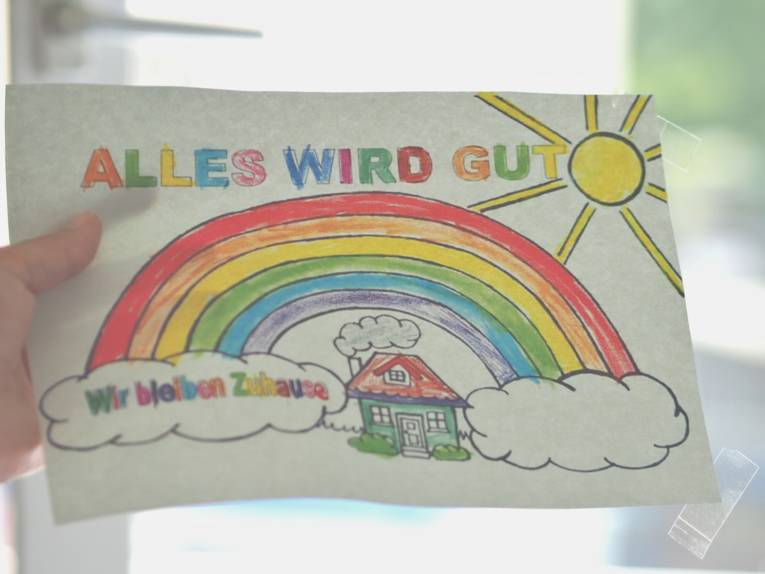 Eine Hand klebt ein von Kinderhand ausgemaltes Bild in ein Fenster. Das Bild zeigt einen Regenbogen über einem Wohnhaus. Dazu der Text: "Alles wird gut" und "Wir bleiben zu Hause". 
