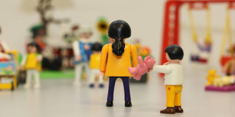 Spielplatzsituation mit Spielzeugfiguren nachgestellt: Ein Kind hebt hinter dem Rücken einer Frau ein Spielzeug.