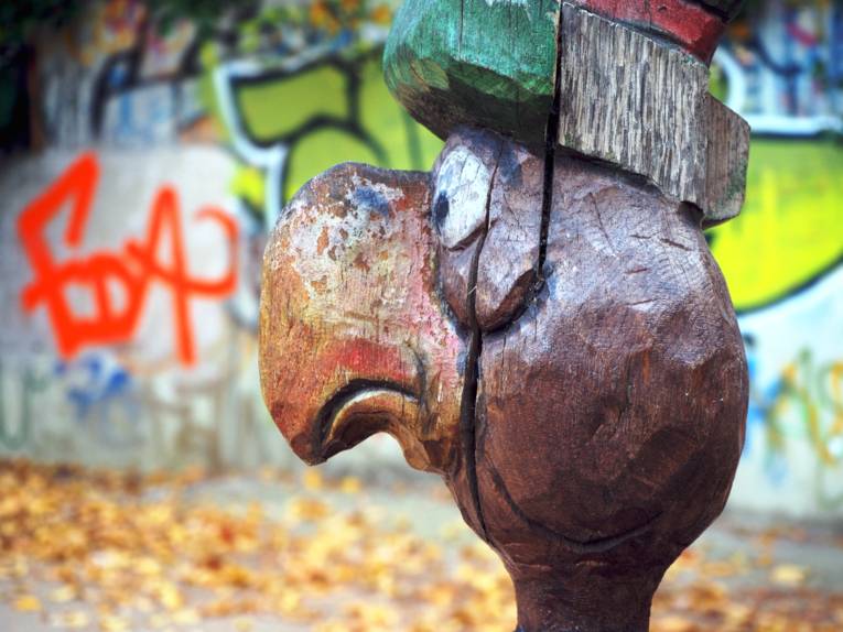 Aus Holz geschnitzter Kopf eines Vogels auf einem Kinderspielplatz, im Hintergrund sind Graffitti.