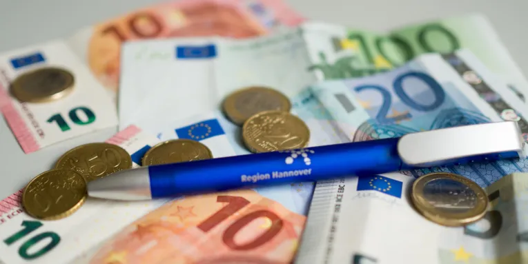 Zwei Zehneuroscheine, ein Hunderteuroschein, ein Zwanzigeuroschein, ein Fünfeuroschein, mehrere Münzen und ein blauer Kugelschreiber liegen auf einem grauen Tisch.
