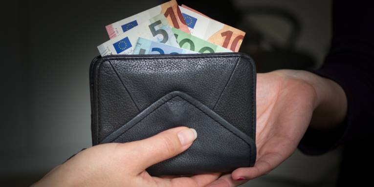 Zu sehen sind zwei Hände von zwei unterschiedlichen Personen und ein schwarzes Portemonnaie mit Eurobanknoten, das überreicht wird.