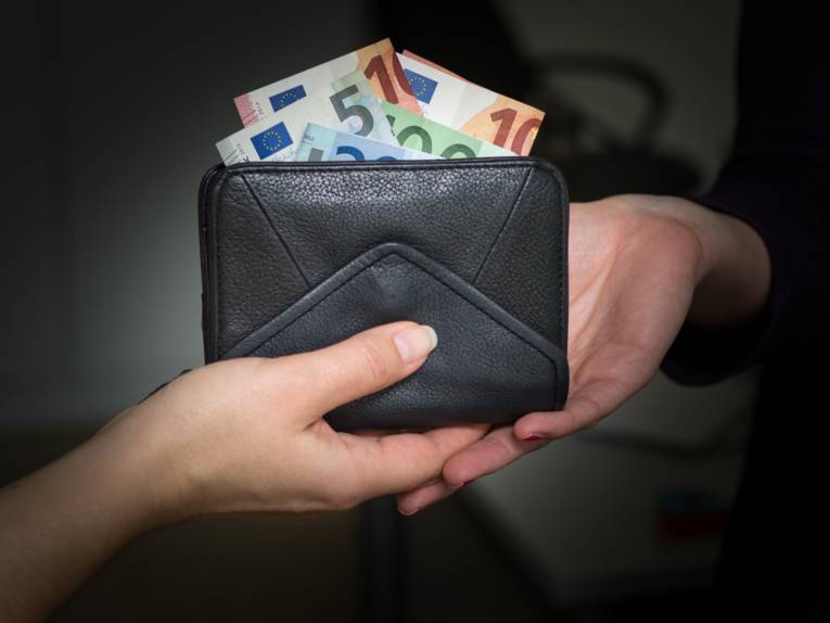 Zu sehen sind zwei Hände von zwei unterschiedlichen Personen und ein schwarzes Portemonnaie mit Eurobanknoten, das überreicht wird.