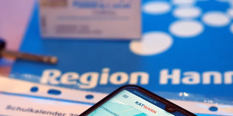 Auf einem Smartphone ist die Katwarn-App geöffnet. Das Smartphone liegt auf einem Schulkalender, drumherum sind ein Autoschlüssel, eine GVH Monatsfahrkarte und das Logo der Region Hannover.