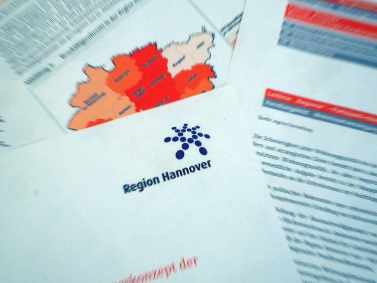 Das Logo der Region Hannover ist auf Papier gedruckt, unter der Papierseite liegen weitere Seiten mit zum Beispiel einer Karte der Region Hannover und ihrer Kommunen und Text.